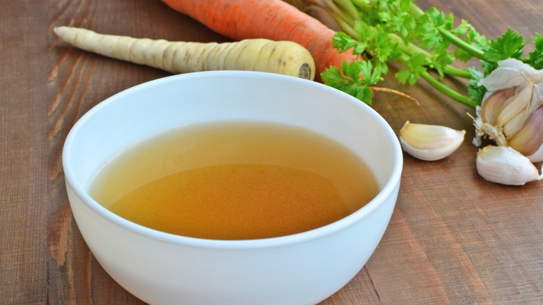 A bowl of beef consomme/broth/etc. in front of garlic, carrots, and parsnip.