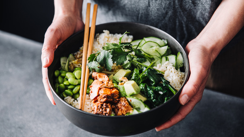 Two hands holding a black bowl of poke with avocado, cilantro, cucumber, and edamame