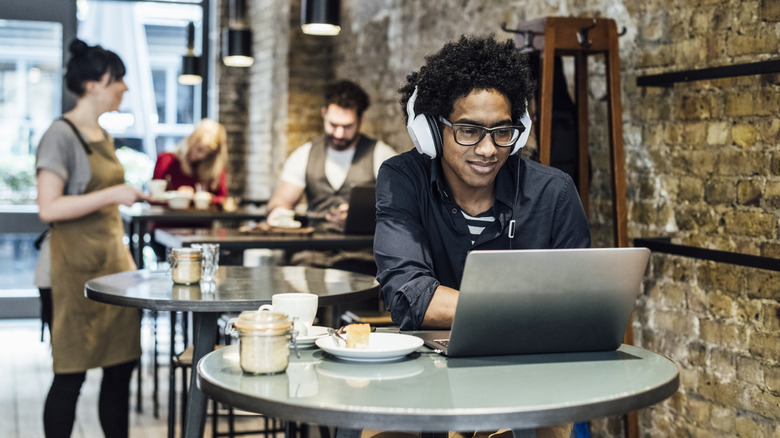 Person sitting at cafe table on laptop with headphones on and other customers behind him