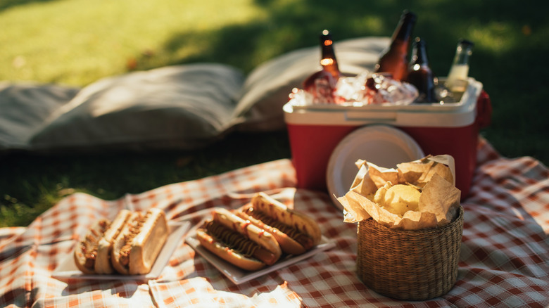 Picnic food on a tablecloth with drinks in a cooler nearby