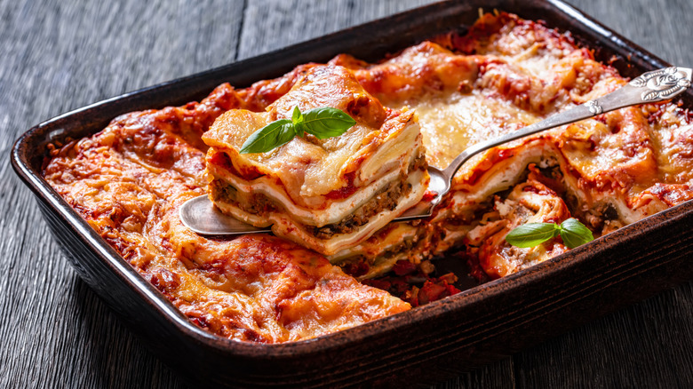 A piece of lasagna sitting on top of a utensil that is in turn sitting on top of a pan of lasagna