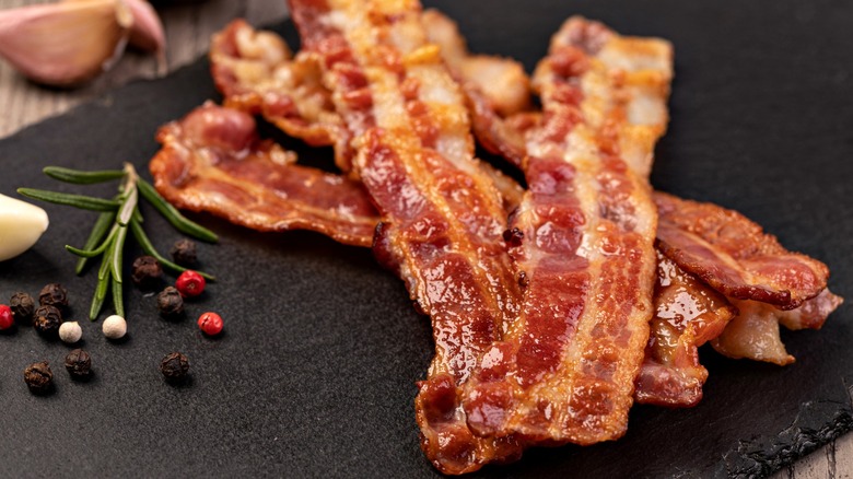Bacon on a black surface with peppercorns and a rosemary sprig.