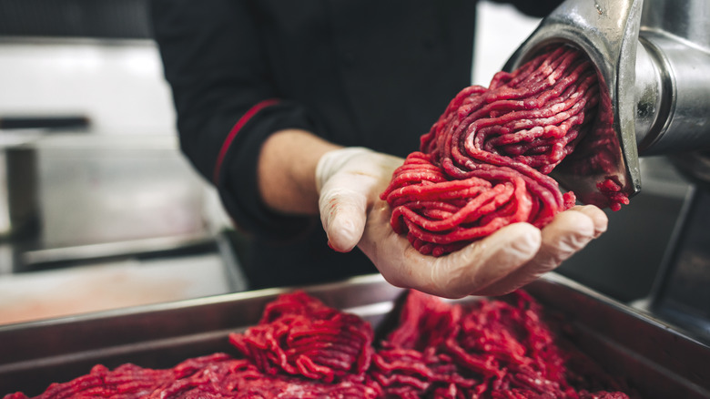 A butcher wearing gloves with freshly ground beef in hand from a large silver meat grinder.