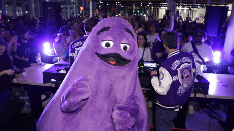Grimace mascot posing in front of a DJ booth at a party.