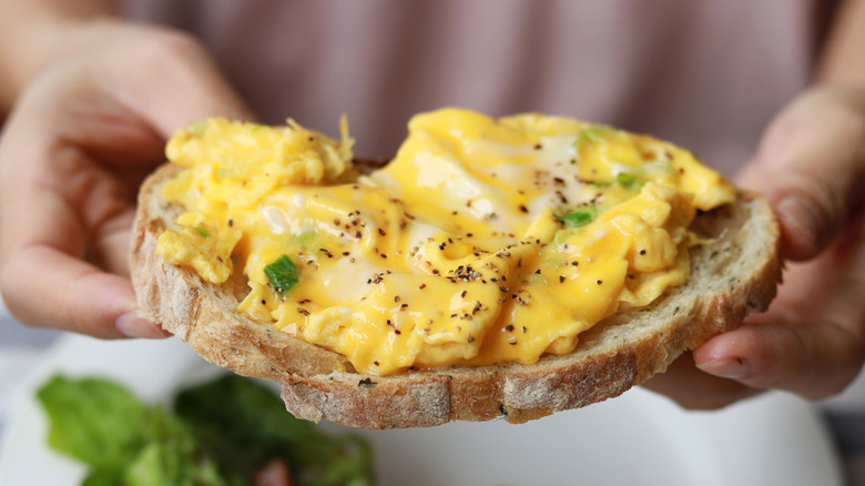 Person holding slice of sourdough topped with scrambled eggs