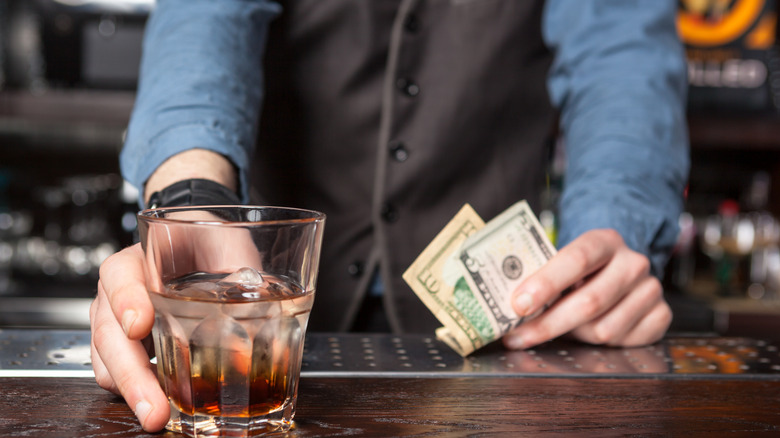A bartender holding $15 in one hand and a drink in the other