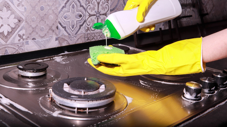 cleaning gas stove top in yellow gloves, dish soap, and sponge