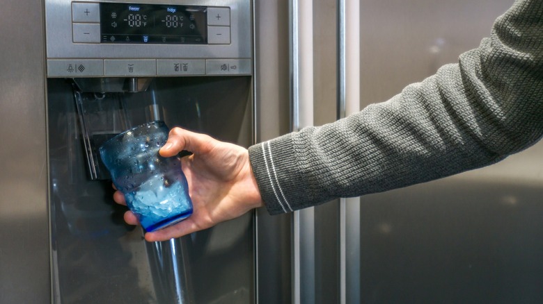 Hand holding a glass with ice under a fridge water dispenser