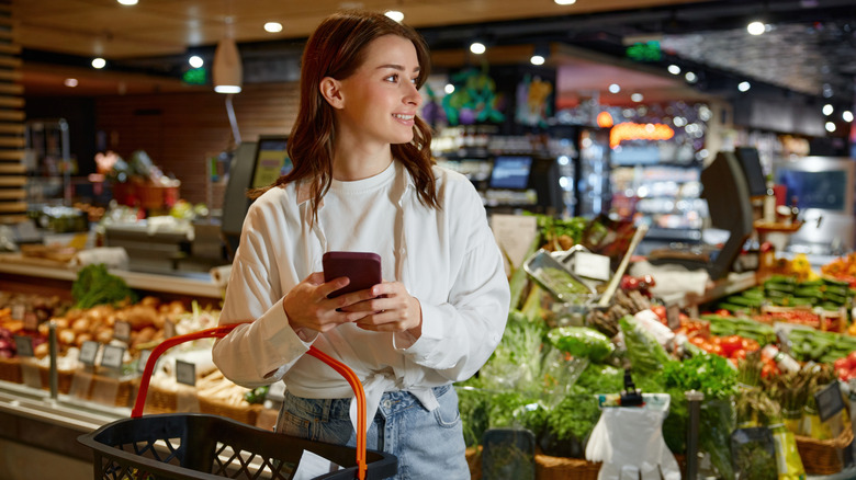 A young woman shopping for groceries checks her phone.