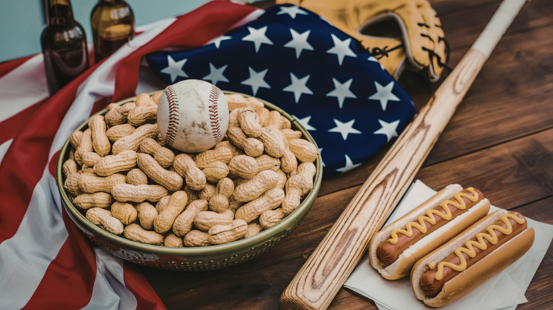 A baseball sitting in a bowl of peanuts next to a baseball bat, two hotdogs, and an American flag