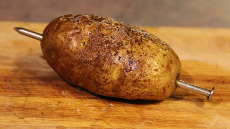 A cooked potato pierced with a metal baking nail on a wood cutting board