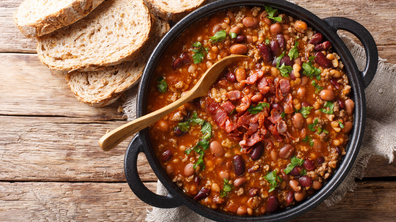 Black pot of bean and meat chili with bacon on top next to sliced bread