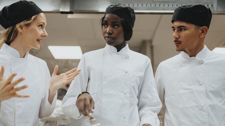 Three chefs in white uniforms and black hats stand in a professional kitchen