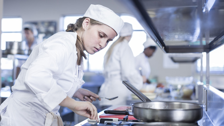 A young chef in a white uniform and cap leans over a stovetop