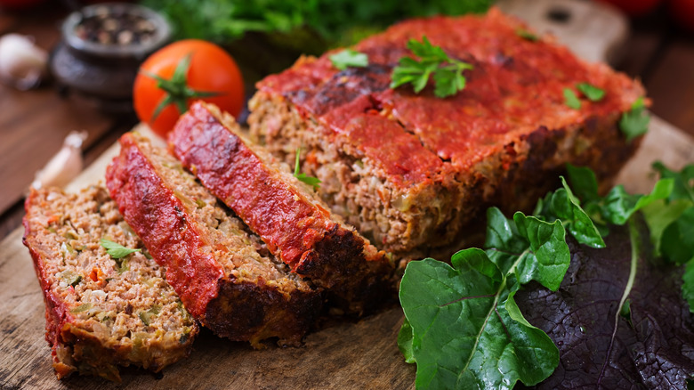 Partially sliced meatloaf with thin tomato topping on a wooden board with herbs and a tomato around it