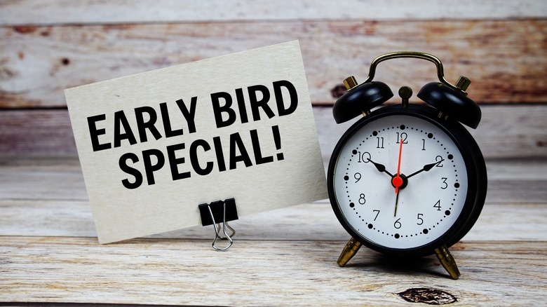 Clip holding up an off-white card with Early Bird Special written on it in black next to a black old-fashioned alarm clock, on a light wood surface