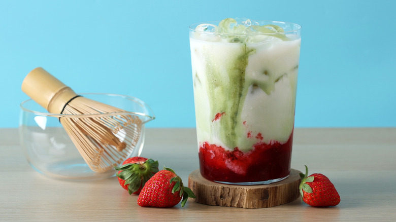 Iced matcha latte with strawberry puree at the bottom of the clear glass with strawberries and a whisk nearby