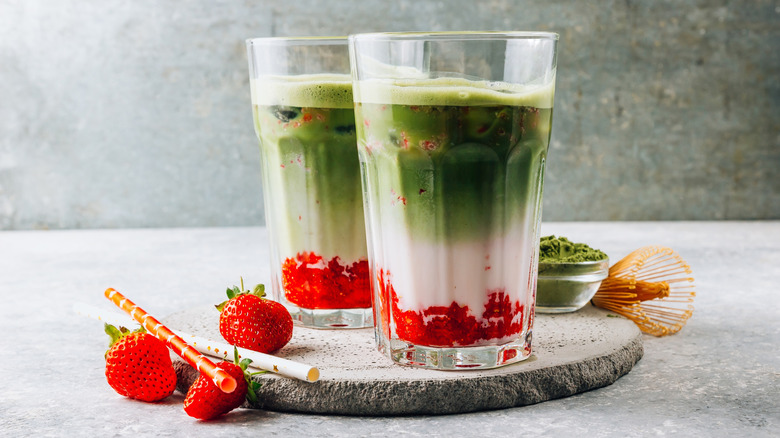Two strawberry matcha lattes in clear glass cups on a stone place setting with strawberries, matcha powder, and a whisk