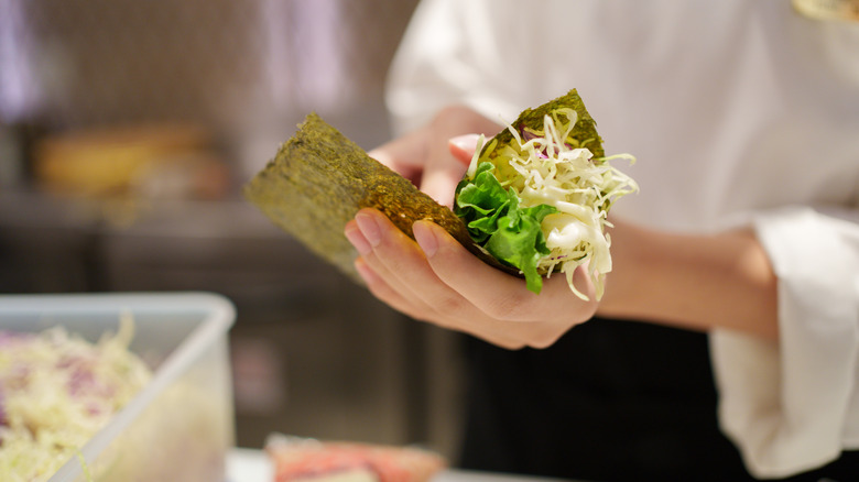 Close-up of a person assembling a temaki hand roll with seaweed, lettuce, shredded cabbage, and mayonnaise. The person is wearing a white chef's jacket, and the background shows a container of vegetables in a kitchen setting.