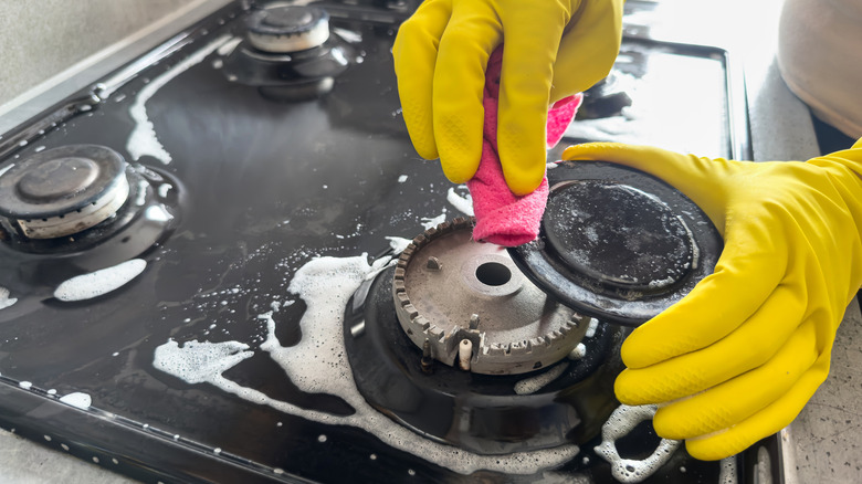 Hands in yellow gloves scrubbing gas stove cooktop