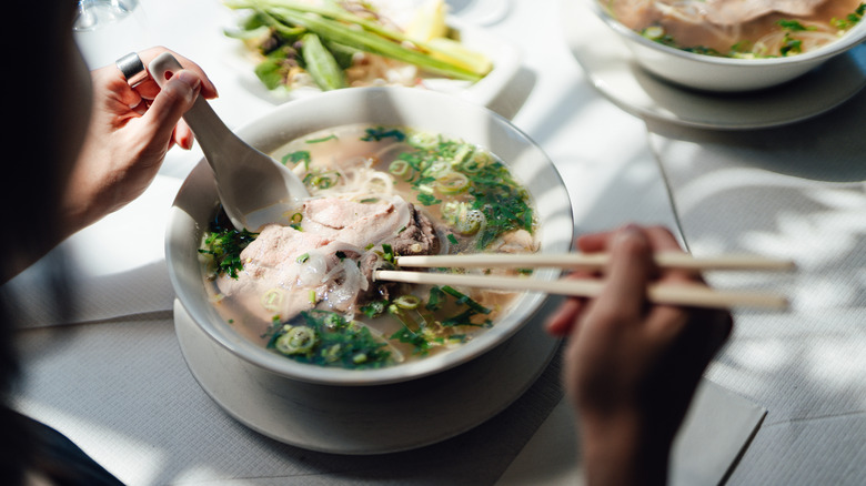 Person eating pho in a restaurant with a spoon and chopsticks