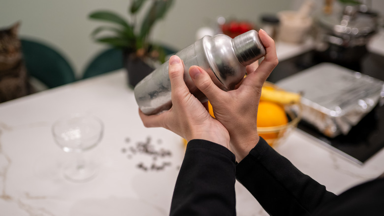 A person using a cocktail shaker over a kitchen Island with a cat nearby.