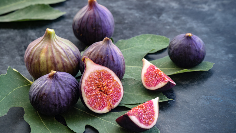 Purple figs on a couple of large green fig leaves with one sliced half on a black surface