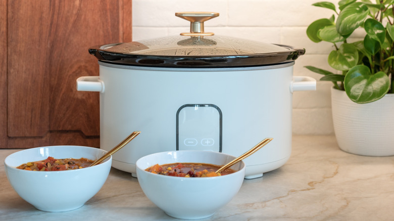 A slow cooker on a kitchen bench with two bowls of beef stew