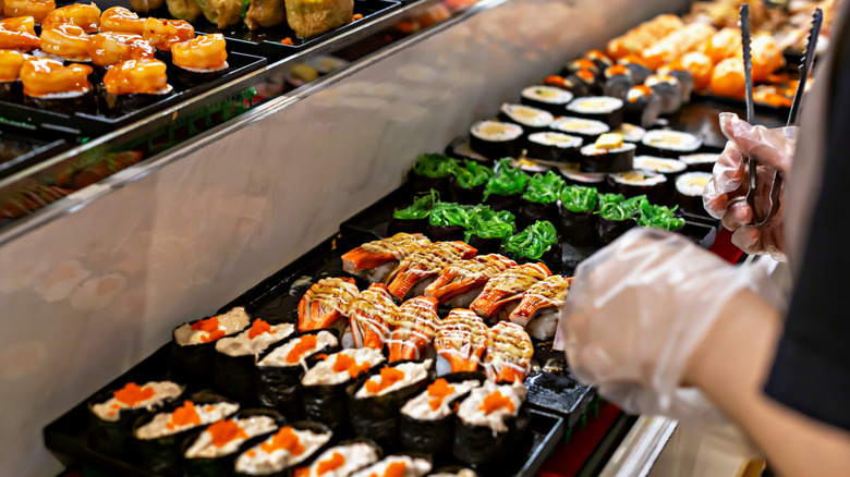 A person holding tongs near several platters of different kinds of rolls at a sushi buffet