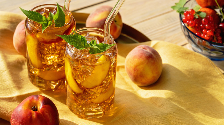 Two glasses of sweet tea with straws and peaches on a light cloth with fruit bowl nearby.