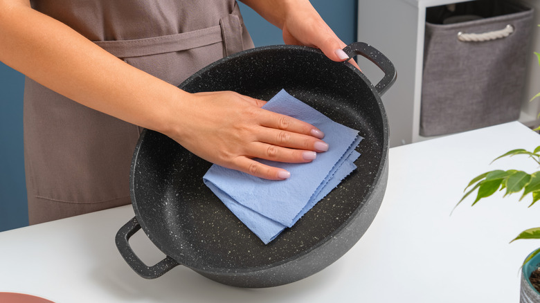 A hand wipes a black cast iron pan with a blue paper towel in the kitchen.