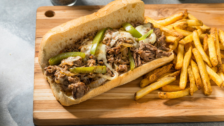 A homemade Philly cheesesteak with onions and peppers on top of it and French fries on the side
