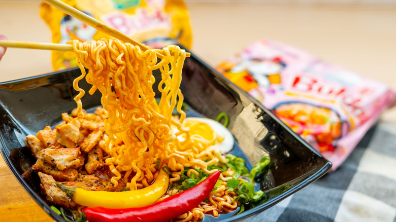 A bowl of spicy Buldak ramen with peppers and egg with packages of Buldak ramen in the background