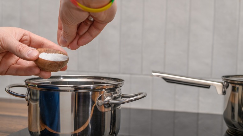 Hands pinching salt from a small wooden bowl to season something cooking in a stainless steel pot on a stove