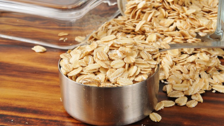 A measuring cup overflowing with raw rolled oats on a wood surface