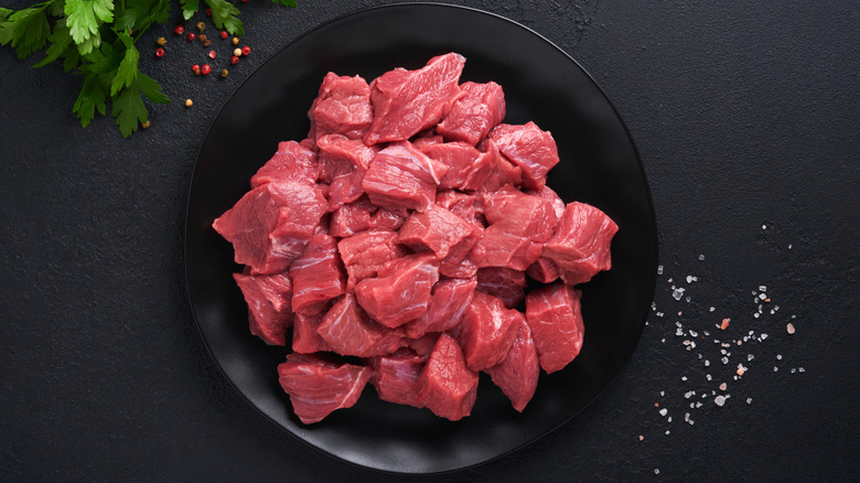 Raw stew meat on a black plate with salt and peppercorns near it.