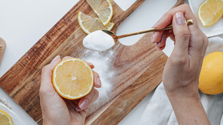 A hand sprinkling baking soda on a wood cutting board and another hand holding a lemon half