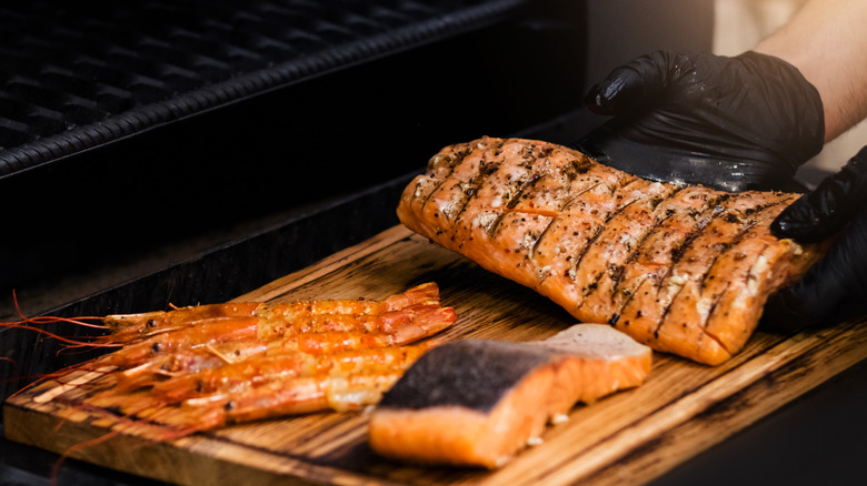 Fish being taken off the grill and placed on a wooden board by a person with black gloves on.
