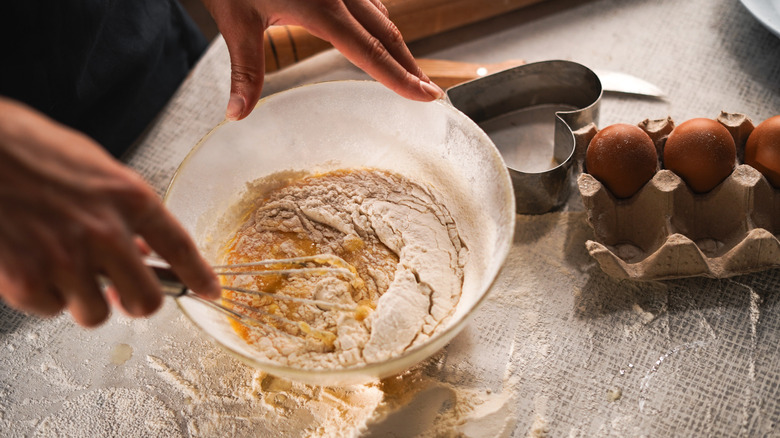 A baker whisks flour and eggs in a bowl on a floured surface with fresh eggs in a carton