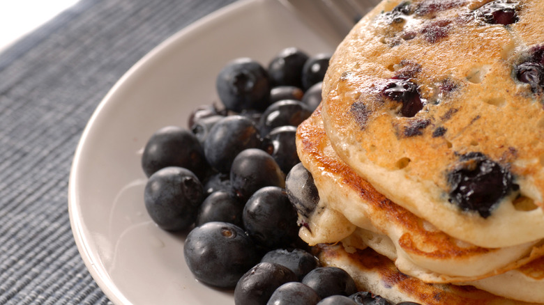 A cropped photo of a white plate of blueberry pancakes with fresh blueberries on the side