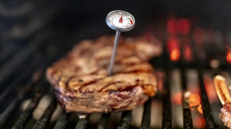 A manual meat thermometer sticks out of a steak on the grill