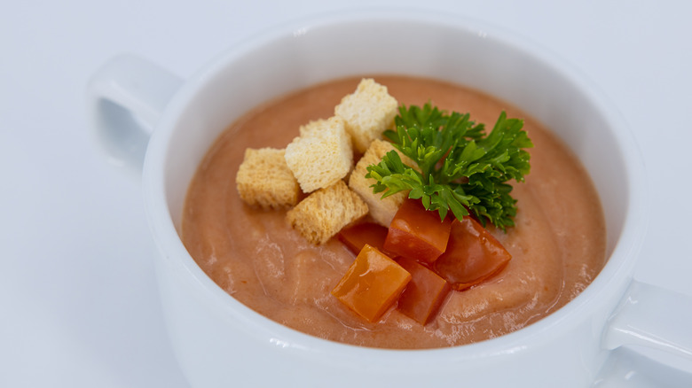 Creamy tomato soup garnished with croutons, parsley, and tomatoes in a white cup.