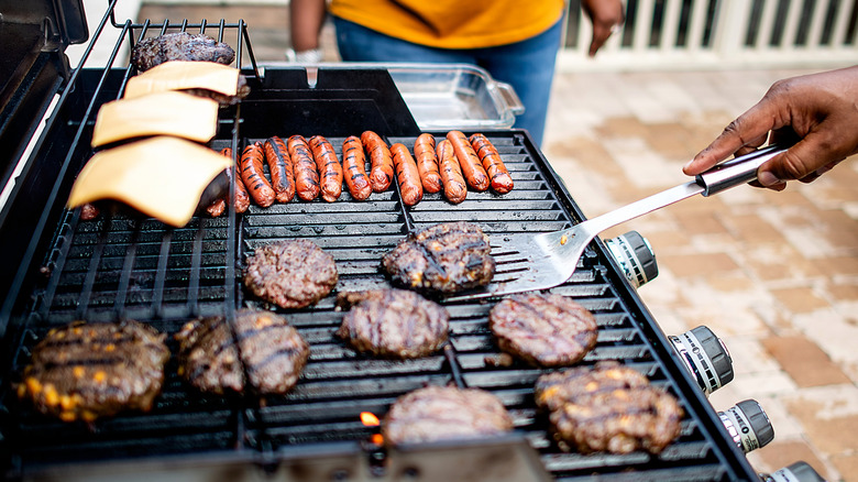 Hamburgers, cheeseburgers, and hot dogs cooking on a grill, with a man holding a spatula under one burger