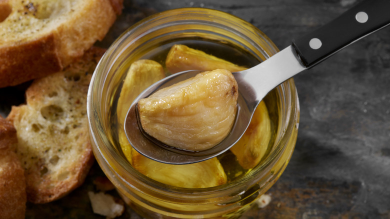 A spoon raises a clove of garlic from a jar of garlic confit near slices of baguette
