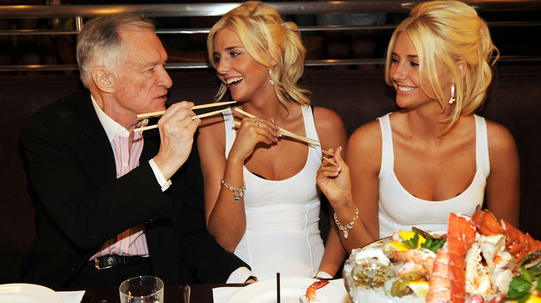 Hugh Hefner holds chopsticks with 2 blonde women in white dresses