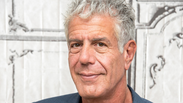 Anthony Bourdain standing in front of a white-painted wall wearing a gray jacket