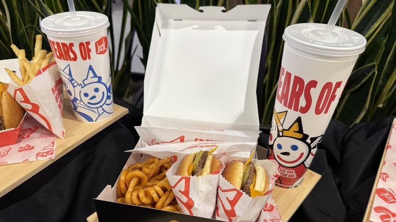 Jack in the Box food on display in takeout box with hamburgers, curly fries, and large takeout drinks