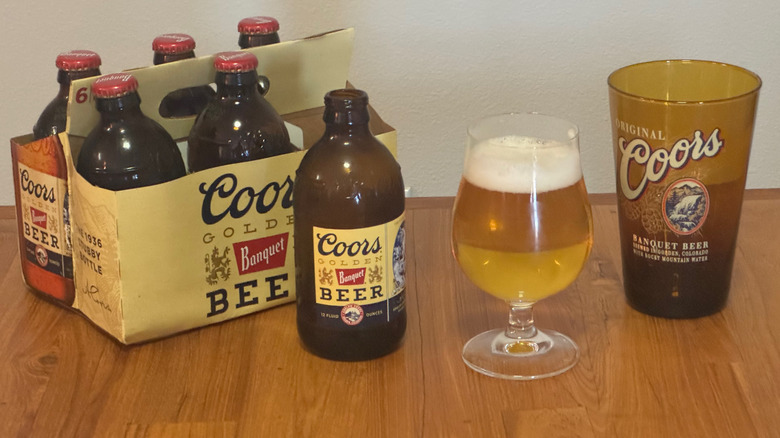 A brown, stubby bottle of Coors on a wooden table next to a six-pack, a full beer glass, and a brown pint glass with the Coors logo painted on it.