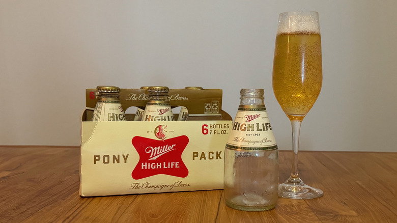 An small, empty glass bottle of Miller High Life next to a six pack of small bottles and a Champagne flute filled with beer.