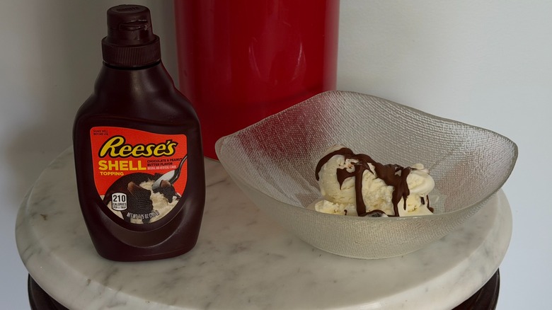 A bottle of Hershey's Reese's chocolate peanut butter shell topping next to bowl of vanilla ice cream with the topping on it